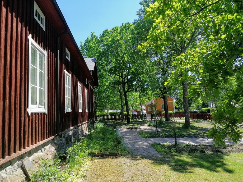 Punainen museorakennus. Piha, punainen aita. Portti auki. Kesä.