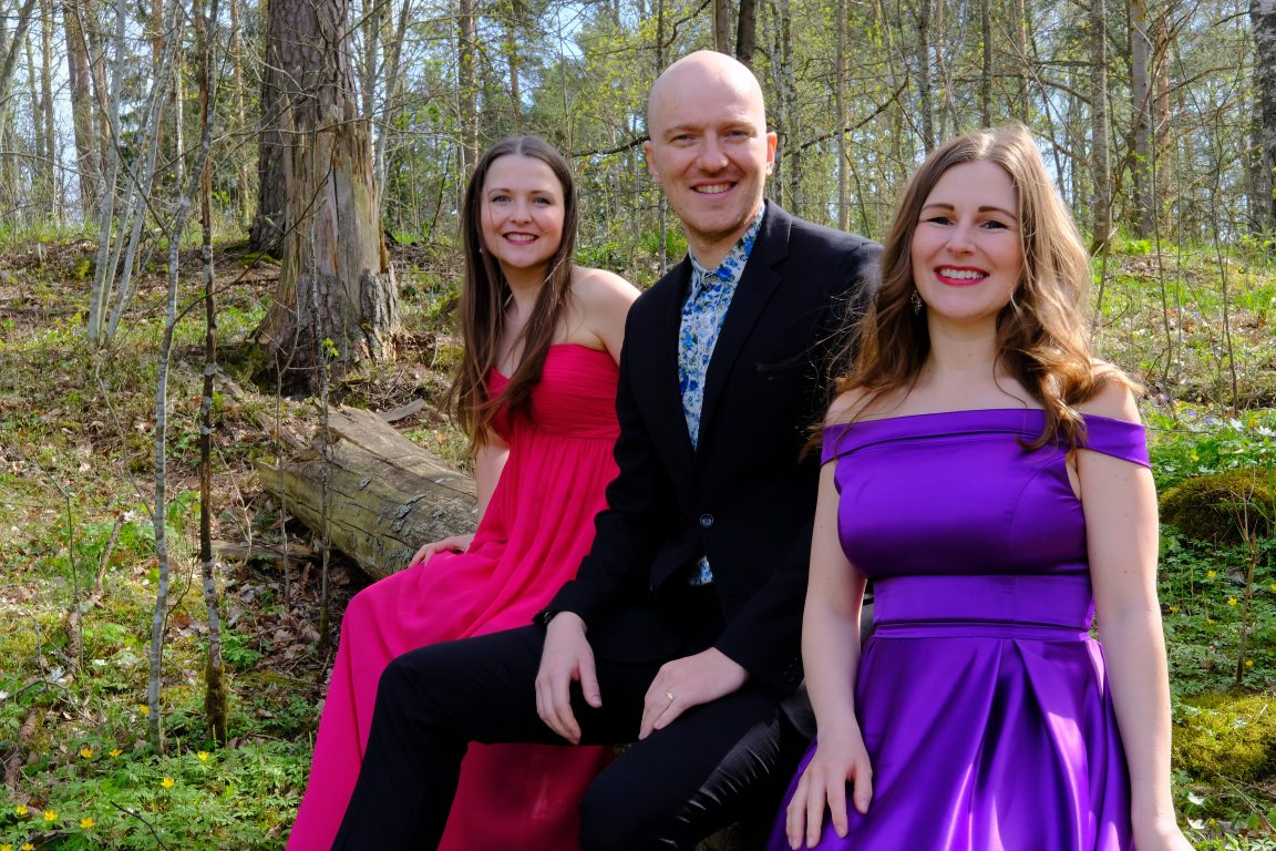 Kolme esiintyjää metsässä, naisilla vaaleanpunainen ja violetti juhlapuku, miehellä tumma puku 