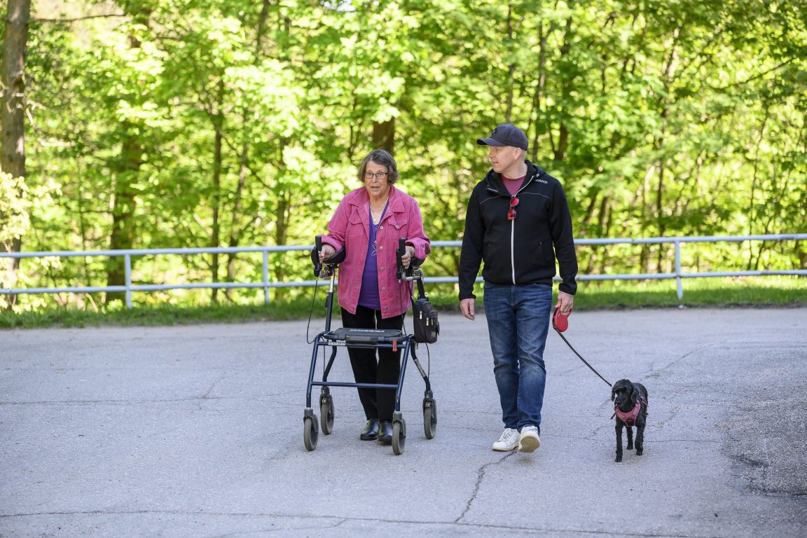 Rollaattoria käyttävä iäkäs nainen kävelee ulkona koiraa taluttavan nuoremman miehen kanssa