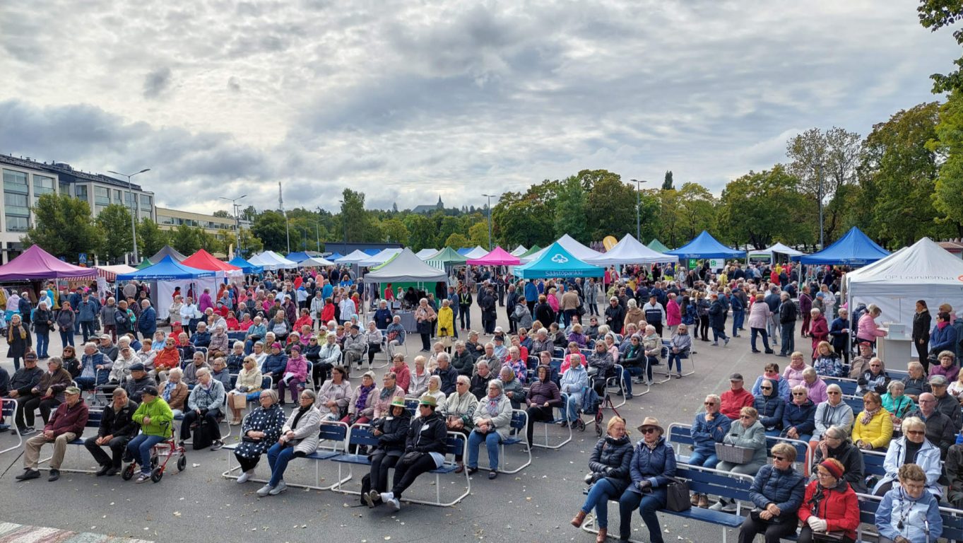 Suuri määrä ihmisiä istuu penkeillä katsomassa torin esiintyjää.
