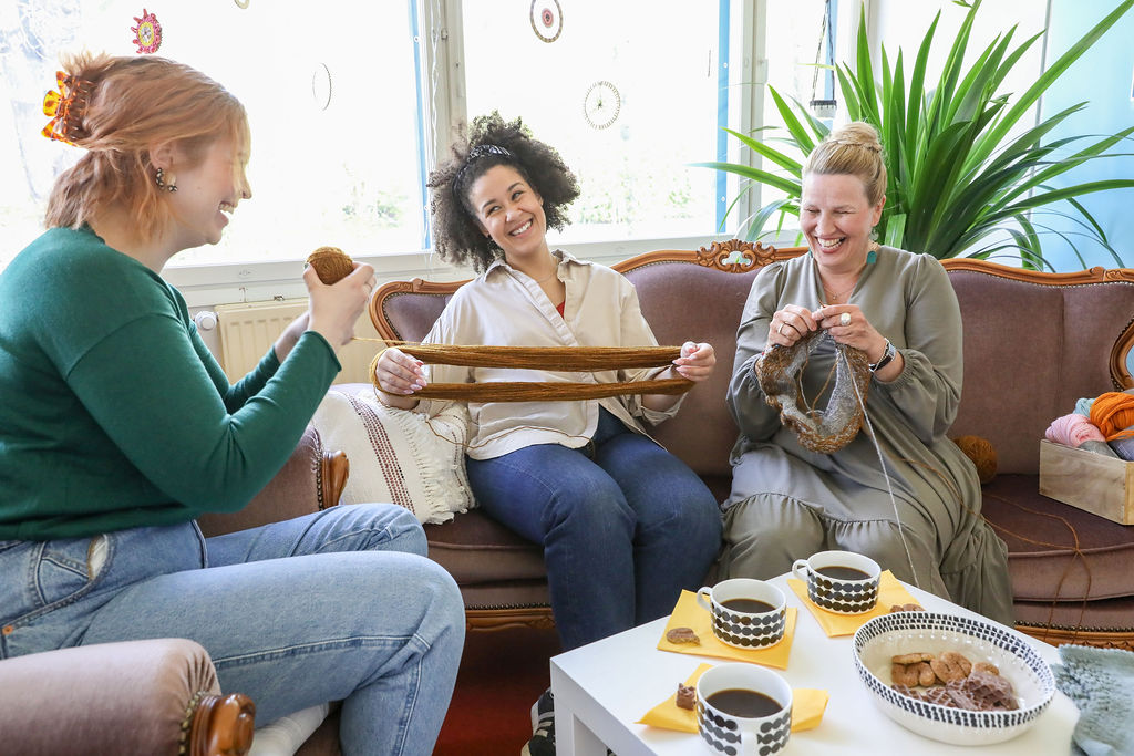Kolme hymyilevää naista istuvat sohvapöydän ääressä kerimässä villalankaa ja kutomassa.