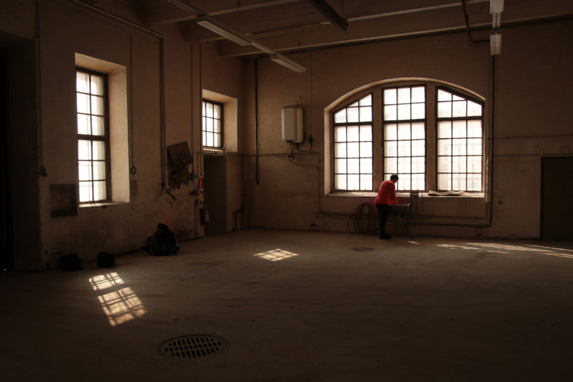 Korjaamo. Tyhjä vanha tehdashalli, jonka pikkuruutuisista ikkunoista paistaa valo tilan lattialle. Yksi henkilö seisoo ikkunan luona.