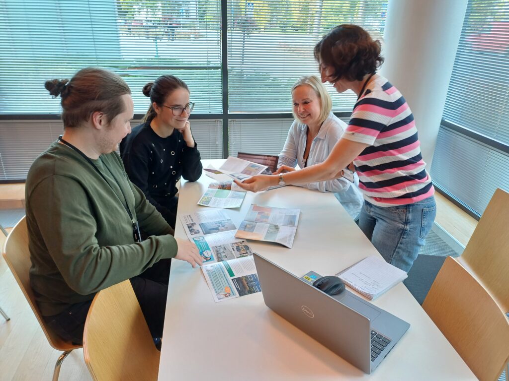 Salon kaupungin maahanmuuttajapalveluiden neljä työntekijää kabinetti Tuomen pöydän ympärillä Salon kaupungintalolla. Pöydällä on esitteitä. Yksi henkilöistä seisoo ja pitää kädessään esitettä. Muut henkilöt istuvat.