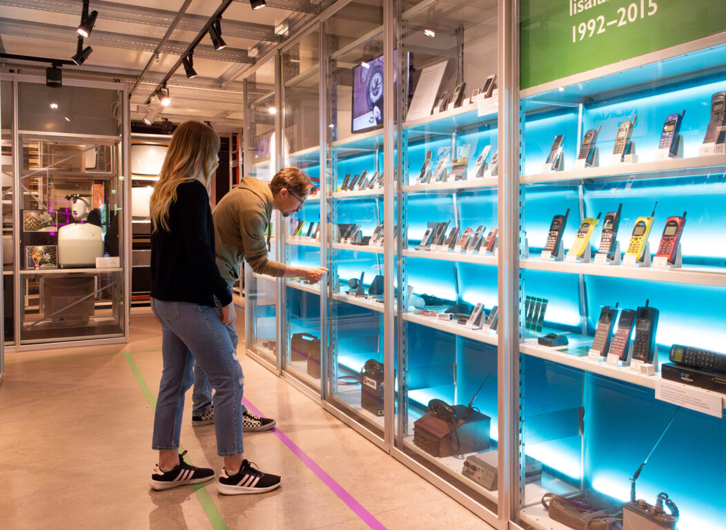 Kaksi museovierasta tarkastelee matkapuhelinvitriiniä elektroniikkamuseon näyttelyssä.