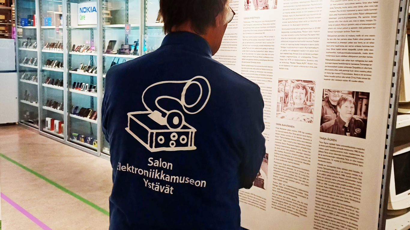 Henkilö Salon Elektroniikkamuseon Ystävät -T-paidassa lukee näyttelytaulun tekstiä.