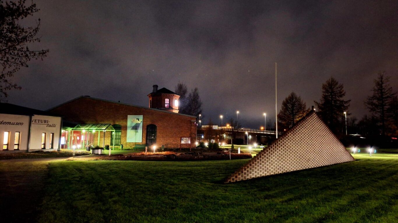 Veturitallin piha-alue, museon sisäänkäynti, valkoinen tiilimuurimainen taideteos ja näyttelyjuliste.