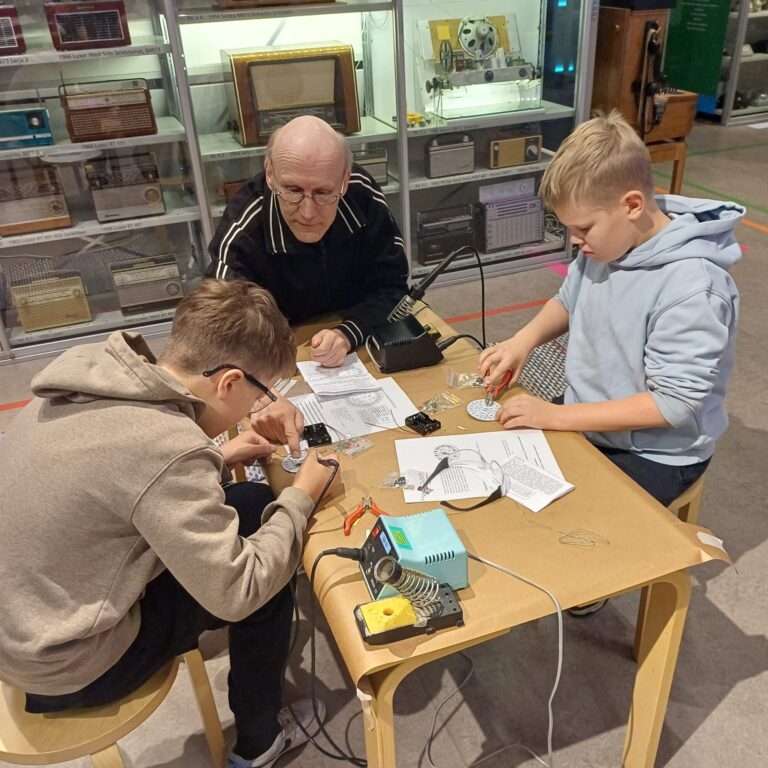 Elektroniikkamuseon työpajassa kaksi poikaa kolvaavat ohjaajan avustuksella.