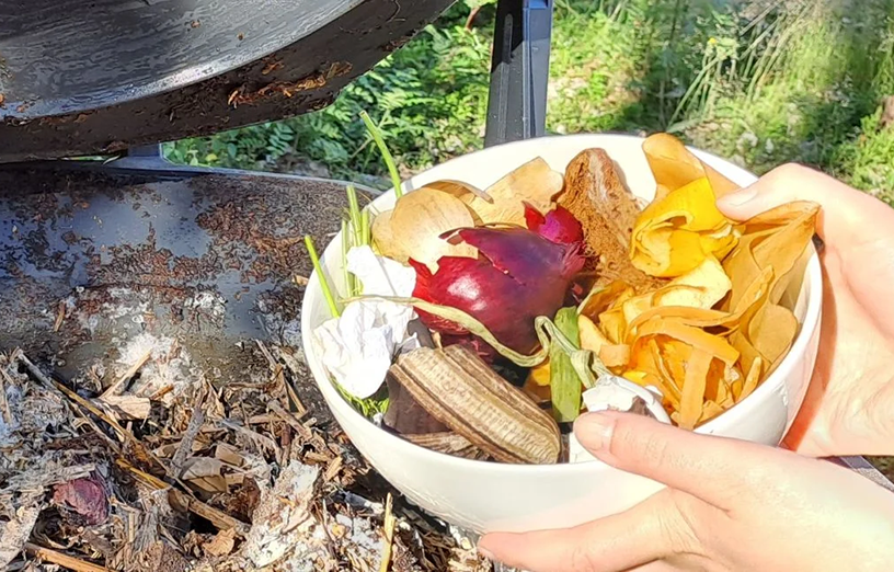 Lähikuvassa käsi, joka laittaa biojätettä avoinna olevaan kompostoriin.