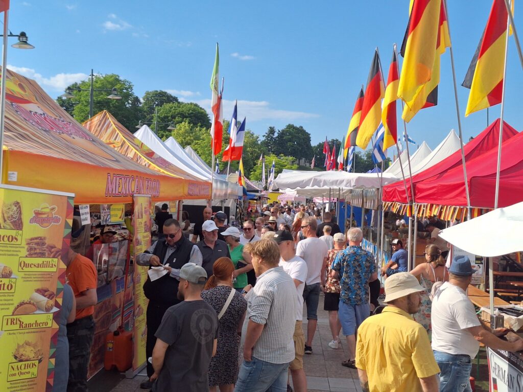 Markkinakojuja, joissa eri maiden lippuja. Ihmisiä kulkee kojujen välissä.