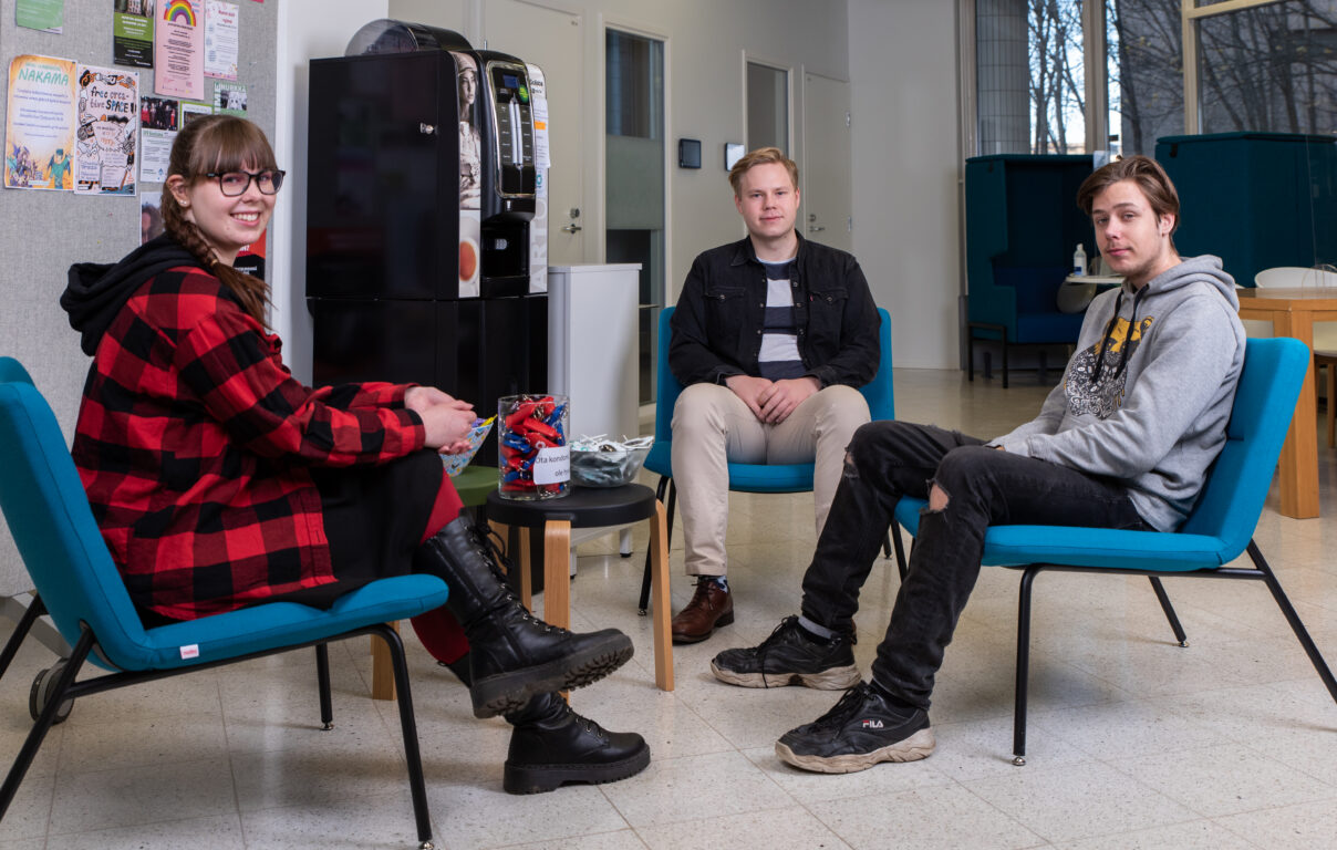 Kolme nuorta istuu tuoleilla pienen pöydän ympärillä ja katsoo kameraan päin.