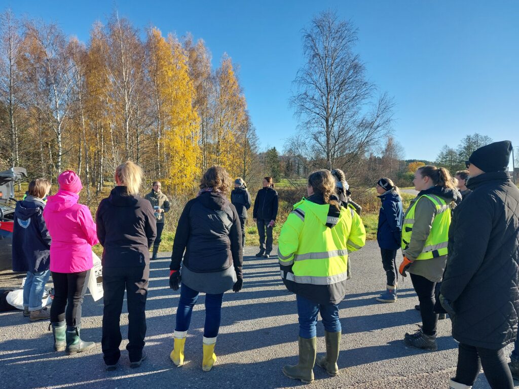 13 henkilöä seisomassa piha-alueella. Yksi henkilöistä puhuu. Muut katsovat puhujaan päin.